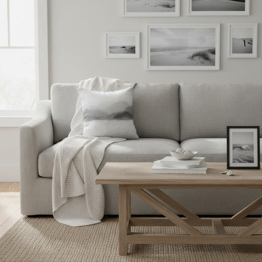 A light gray upholstered sofa in a bright, modern living room, styled with a white throw blanket, a matching pillow, a light wood coffee table, and framed black-and-white wall art.
