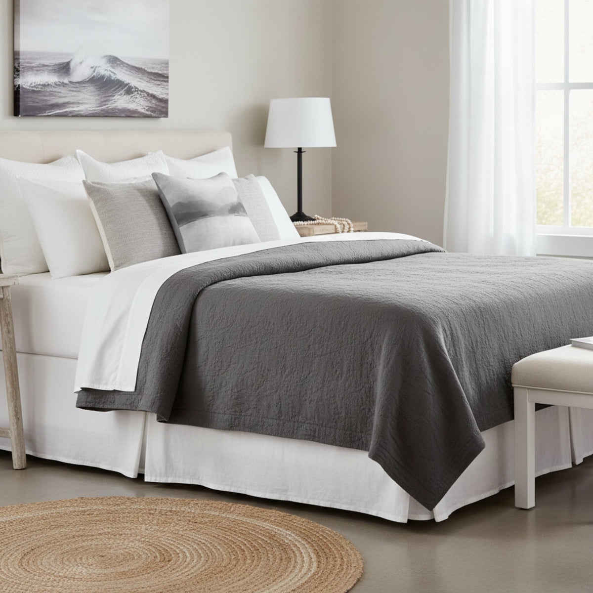 A neatly made bed in a bright bedroom setting, featuring a dark gray textured quilt or comforter layered over white bedding, with matching white and gray pillows. A white table lamp sits on a side table next to the bed, and a framed wave print hangs on the wall above the headboard.