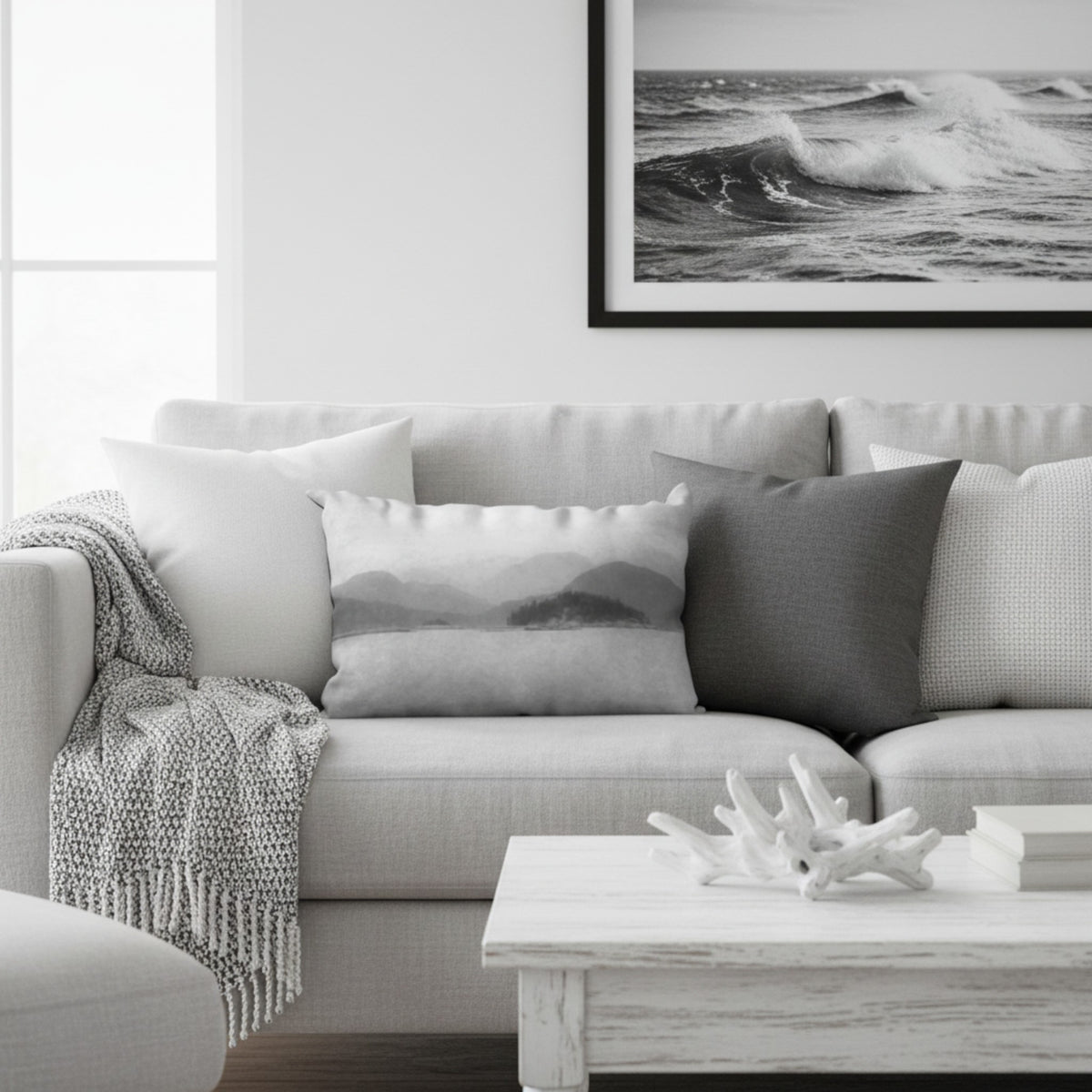 A modern living room scene featuring a light gray sofa styled with a black and white mountain landscape throw pillow, a dark gray pillow, a white pillow, a textured throw blanket, and a framed black and white ocean wave print on the wall.