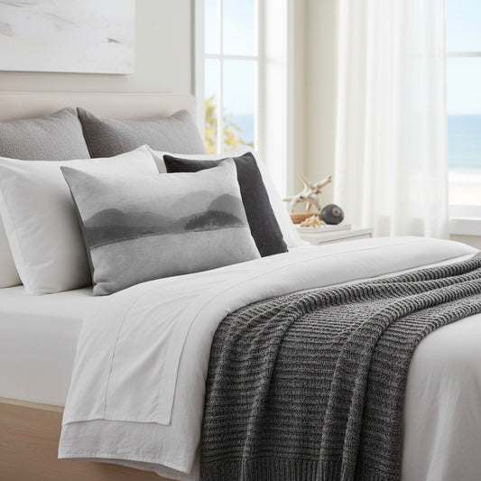 A styled bed in a bright bedroom setting with white bedding, multiple gray and black pillows, a gray knit throw blanket, and a window with sheer curtains and a view of the ocean.