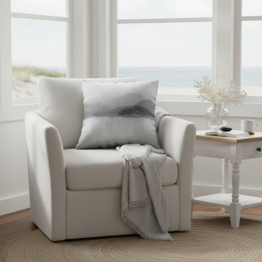 A light beige upholstered armchair in a bright room with large windows overlooking the ocean, styled with two gray pillows, a gray throw blanket, and a small side table nearby.