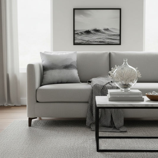 A modern living room scene featuring a light gray fabric sofa with a matching gray throw pillow, a black-framed black and white ocean wave print hanging on the wall, and a white marble-top coffee table in the foreground.