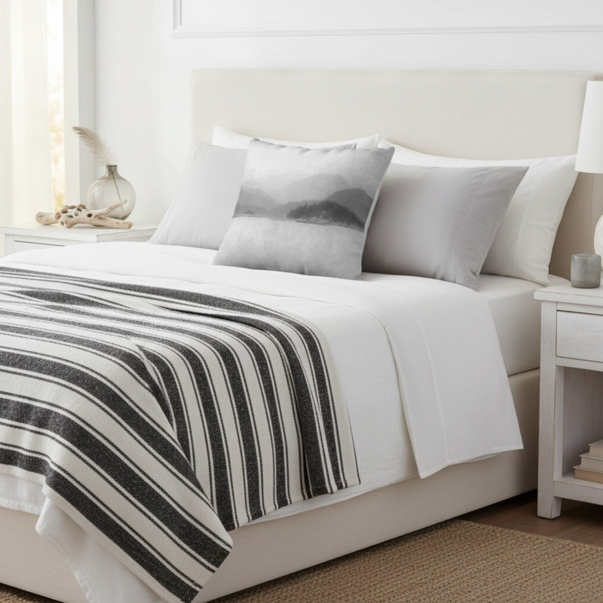 A neatly made bed in a bright bedroom setting, featuring white bedding, a black-and-white striped throw blanket, and a gray mountain-patterned accent pillow.