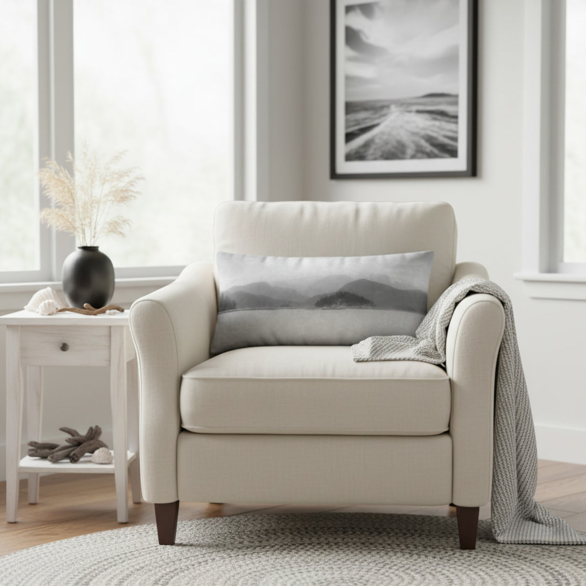 A cream upholstered armchair in a bright living room, styled with a gray mountain-patterned throw pillow, a gray knit throw blanket, and a small white side table beside it.