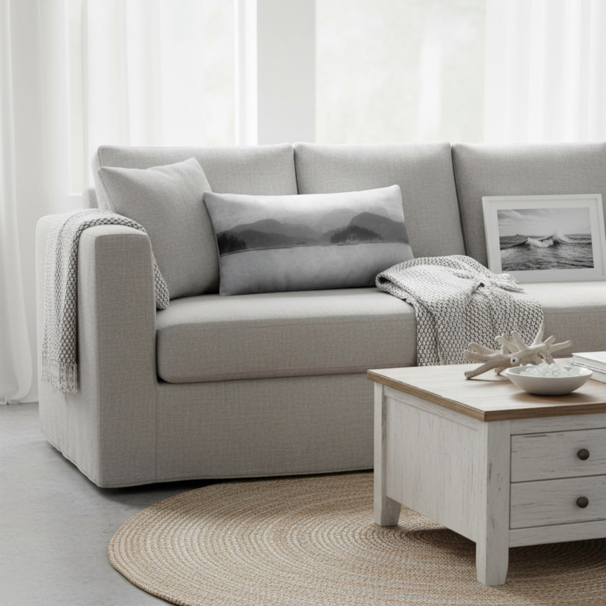 A light gray upholstered sofa in a bright, modern living room setting, styled with a matching gray mountain-patterned pillow, a white knit throw blanket, and a small framed black-and-white picture.