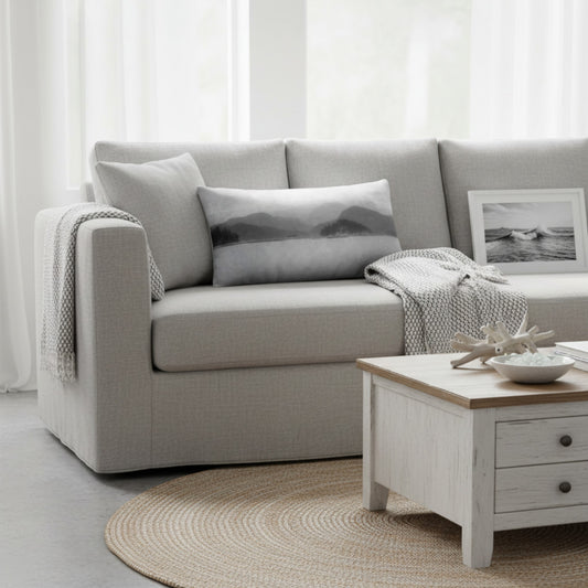 A light gray upholstered sofa in a bright, modern living room setting, styled with a matching gray mountain-patterned pillow, a white knit throw blanket, and a small framed black-and-white picture.
