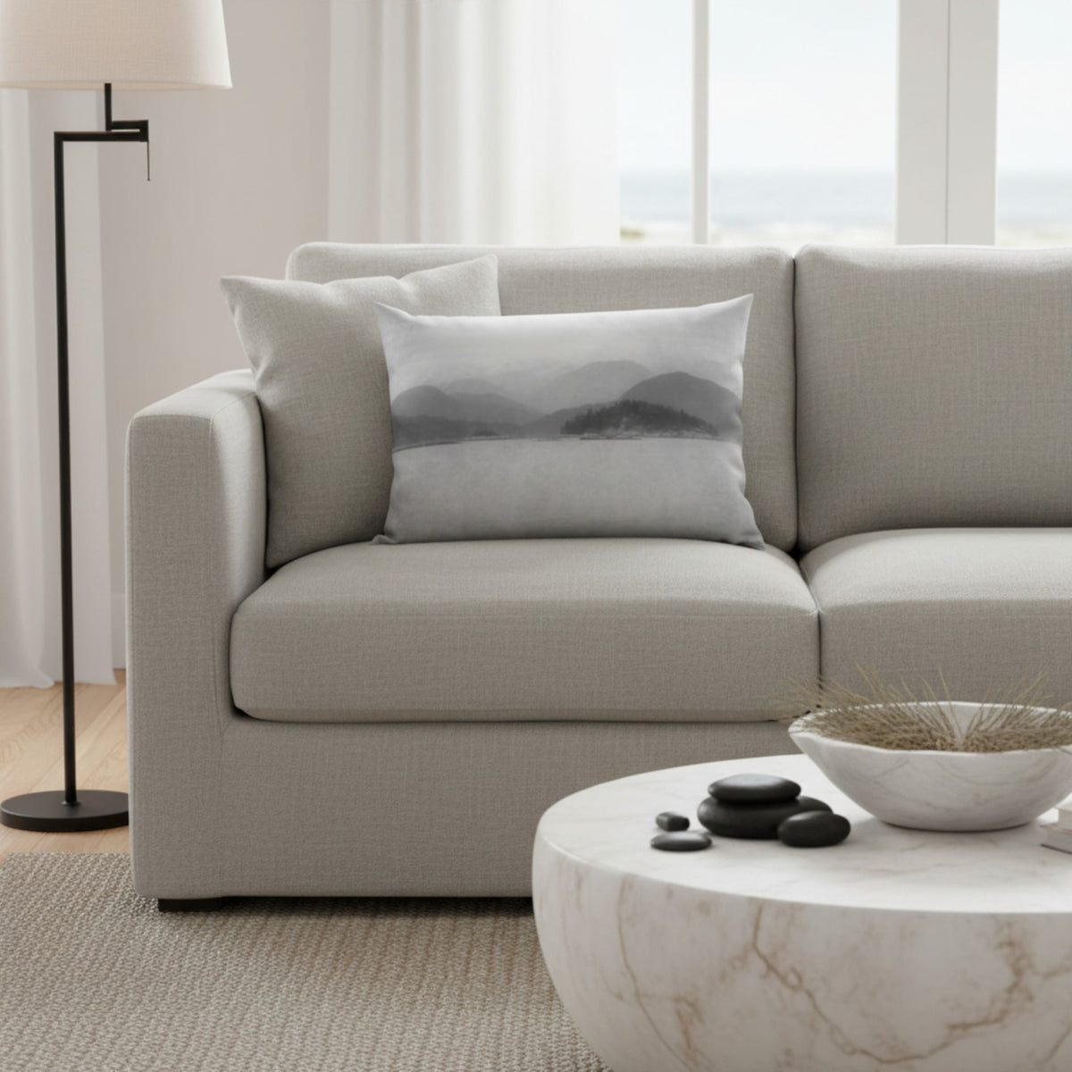 A light gray textured throw pillow with a subtle gray mountain landscape print, styled on a light beige fabric sofa in a bright, modern living room.