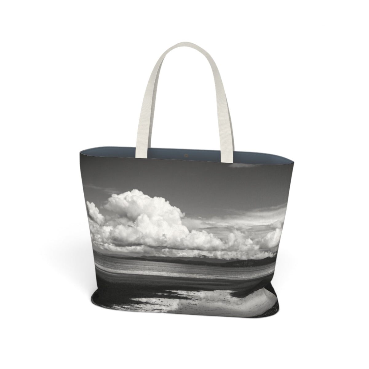 A tote bag featuring a black and white landscape photograph of clouds over water, with light beige fabric handles, shown on a plain white background.