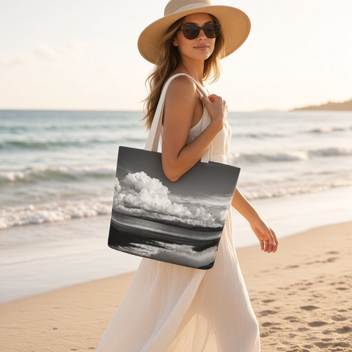 A woman in a white dress and straw hat walks on a sandy beach carrying a large tote bag printed with a black-and-white landscape scene of dramatic clouds over water.