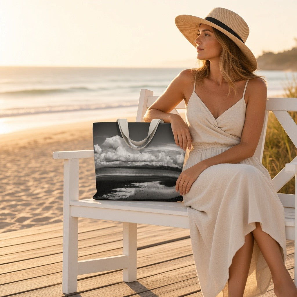 A woman in a cream dress and straw hat sits on a white beach bench at sunset, holding a large tote bag printed with a black-and-white ocean scene. The background shows a sandy beach and ocean.