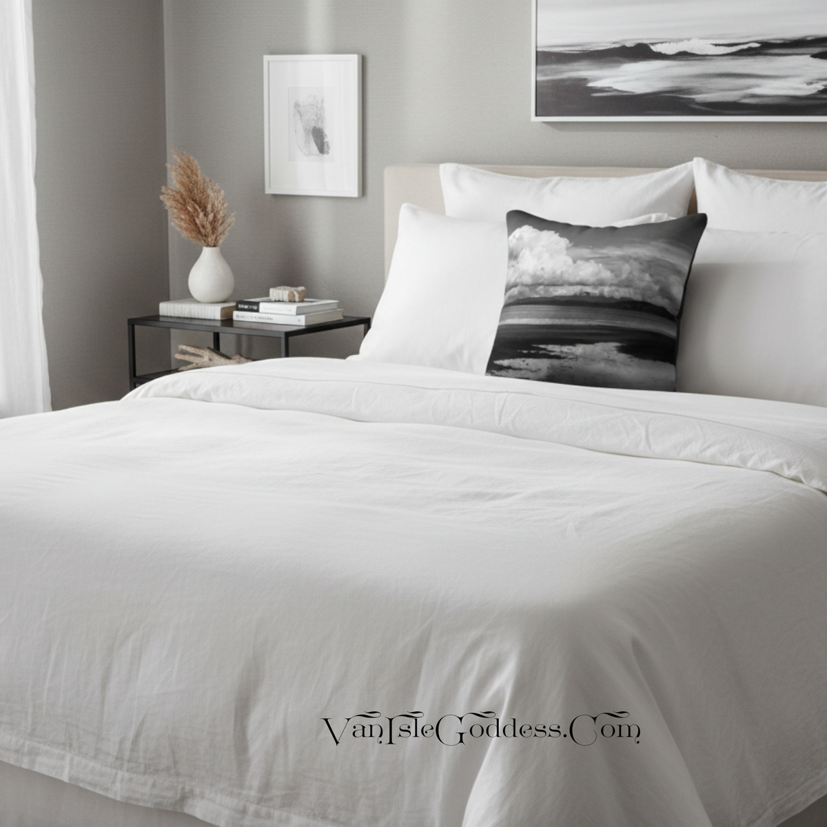 A neatly made bed with white bedding and a black-and-white landscape throw pillow in a styled bedroom. A side table with books and a vase, wall art, and a window curtain are visible in the background.