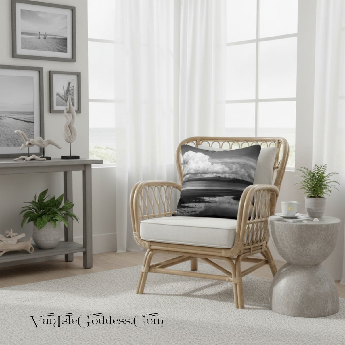 A light natural rattan armchair with a white upholstered seat cushion and a black-and-white landscape throw pillow, styled in a bright coastal-themed living room next to a concrete side table and white sheer curtains.