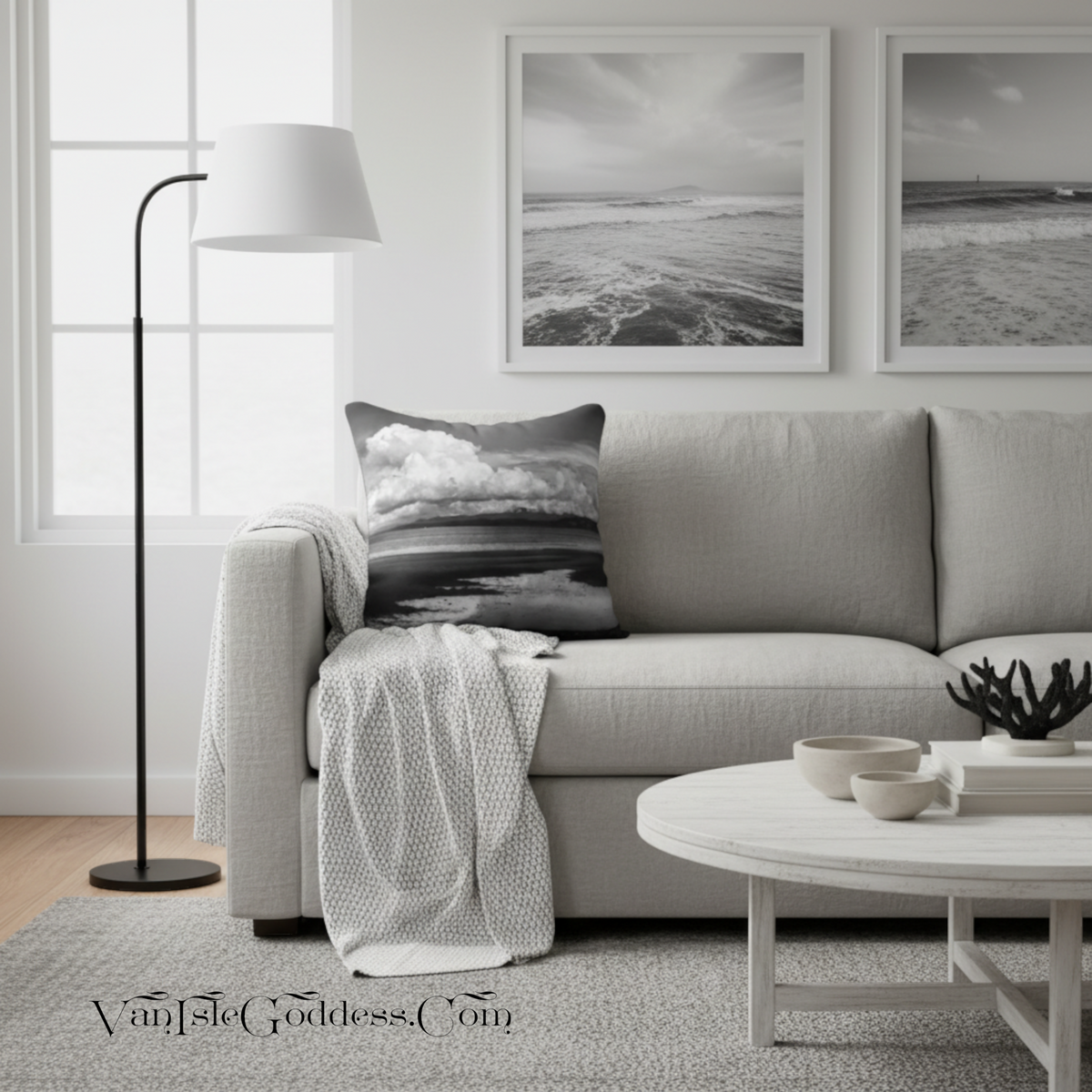 A styled living room scene featuring a light gray sofa with a black-and-white beach print throw pillow, a white throw blanket, a black floor lamp, a round white coffee table, and two framed black-and-white ocean prints on the wall.