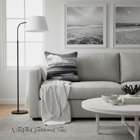 A styled living room scene featuring a light gray sofa with a black-and-white beach print throw pillow, a white throw blanket, a black floor lamp, a round white coffee table, and two framed black-and-white ocean prints on the wall.