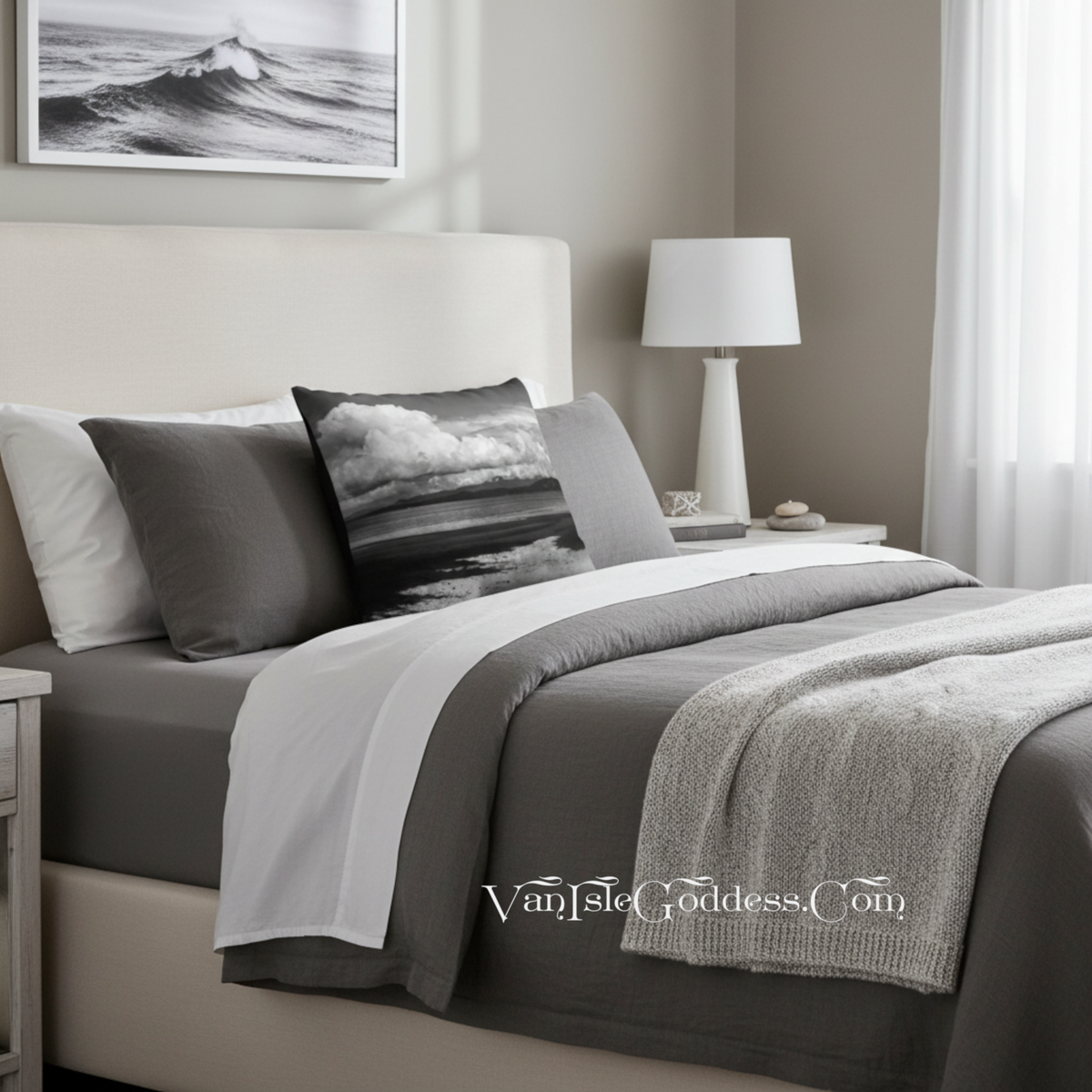 A styled bed in a neutral bedroom setting, featuring a gray and white bedding set with a decorative pillow showing a black and white ocean wave print. The room includes a beige upholstered headboard, a white table lamp, and a framed wave print on the wall.