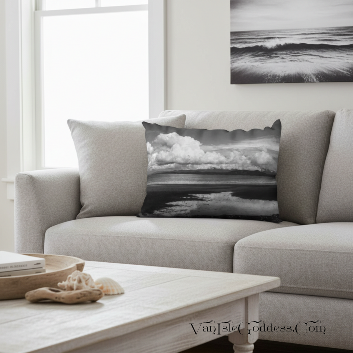 A square throw pillow featuring a black and white ocean landscape print with dramatic clouds and water reflections, styled on a light gray sofa in a bright living room.