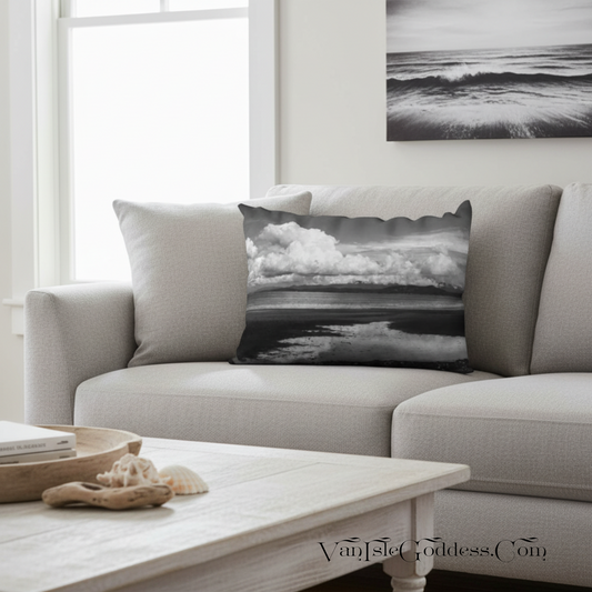 A square throw pillow featuring a black and white ocean landscape print with dramatic clouds and water reflections, styled on a light gray sofa in a bright living room.
