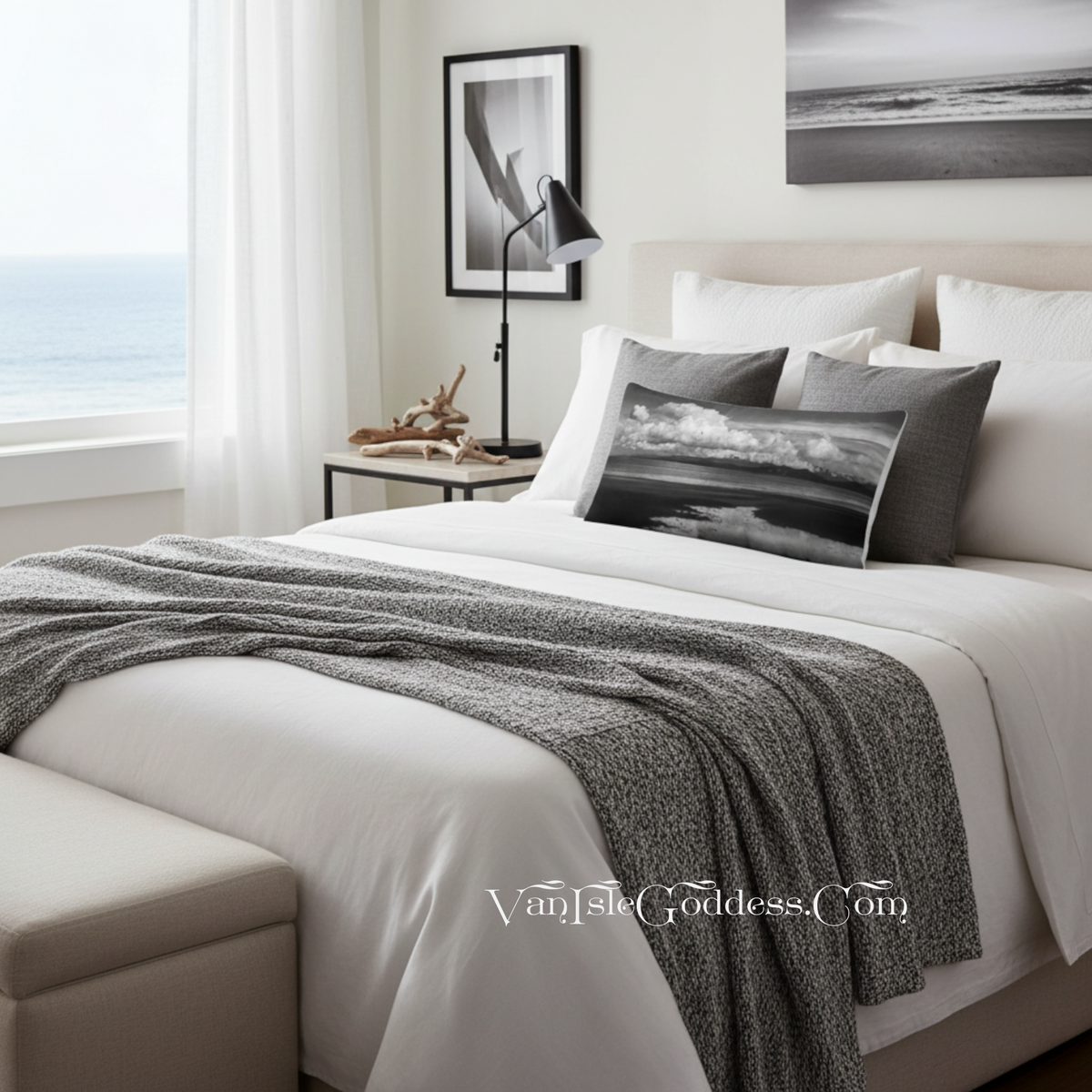 A styled bedroom scene with a bed dressed in white bedding and a textured gray throw blanket, accented by two dark gray pillows featuring a black-and-white seascape print. A window with sheer curtains reveals the ocean outside, and wall art hangs above the bed.