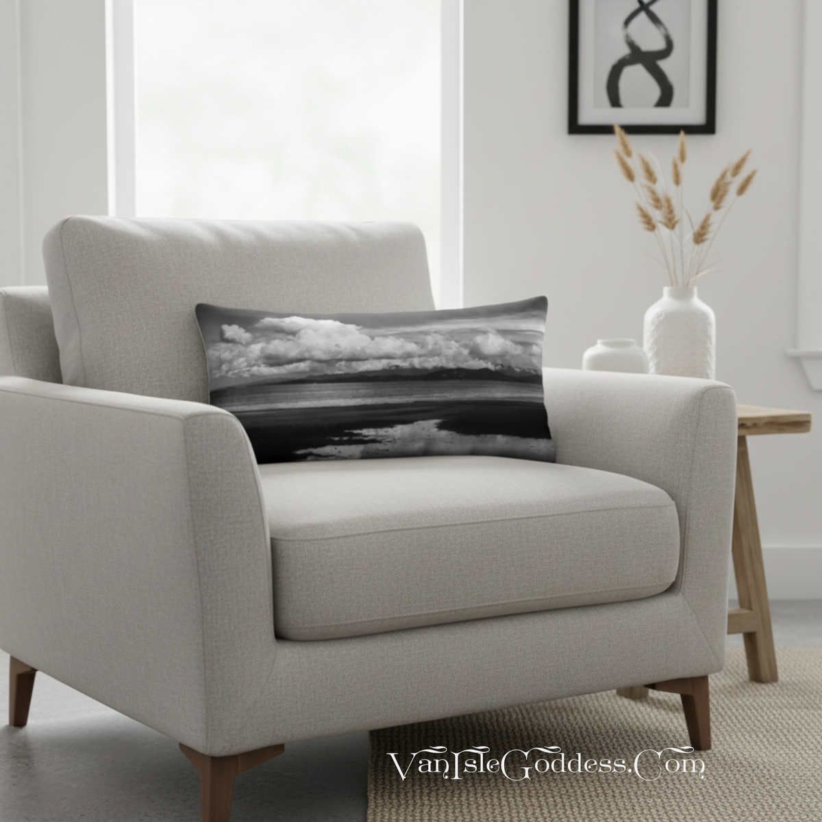 A rectangular lumbar throw pillow with a black and white landscape photograph of clouds and water, resting on a light beige armchair in a modern living room setting.
