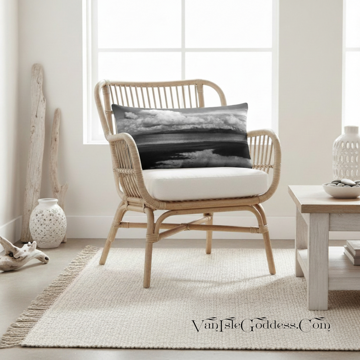 A natural rattan armchair with a white cushion sits on a woven rug in a bright, neutral room. A square black-and-white landscape throw pillow rests on the chair, and a small side table, decorative vase, and driftwood decor are nearby.