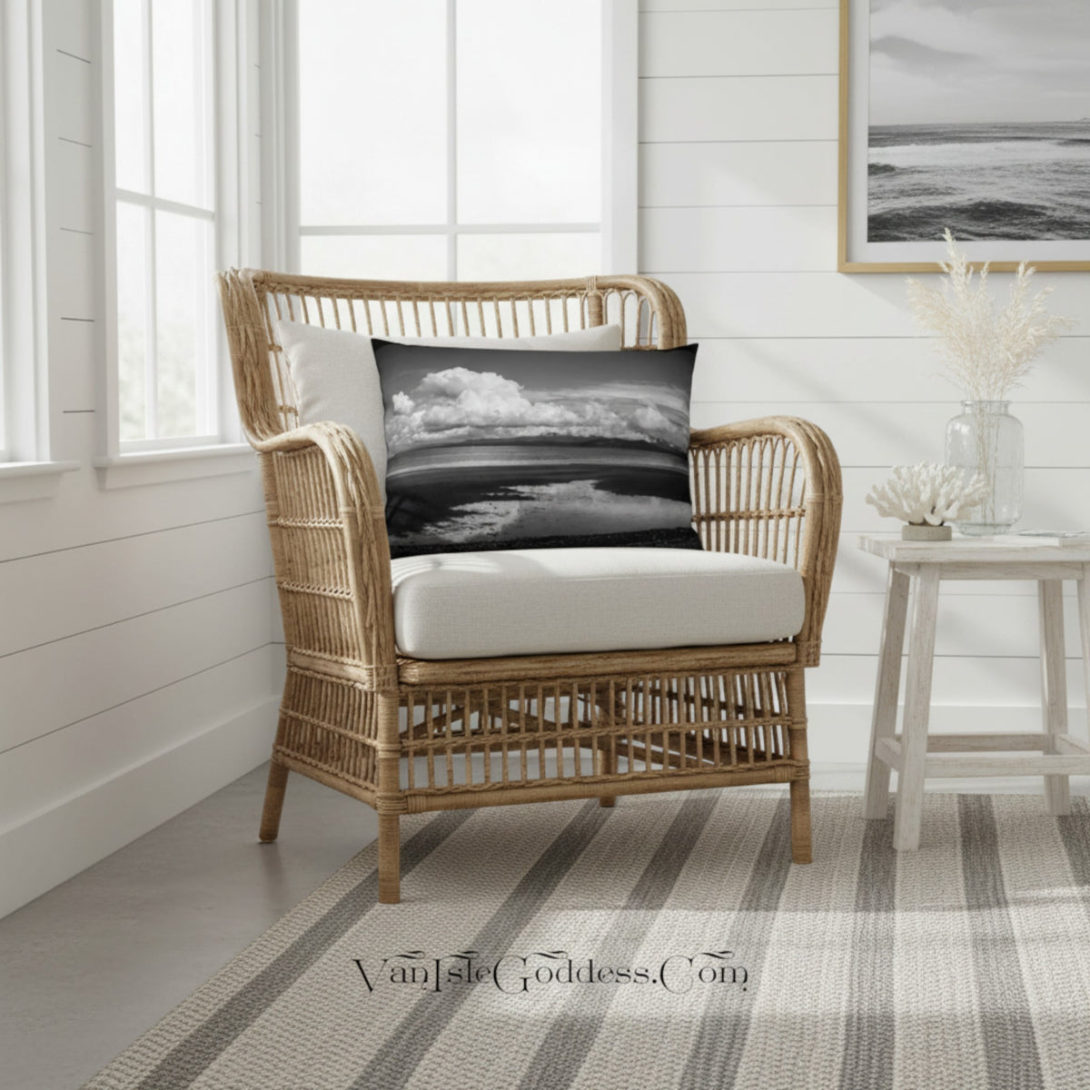 A natural wicker armchair with a white cushion sits in a bright, white-shiplap room. A square black-and-white ocean landscape throw pillow is placed on the chair.