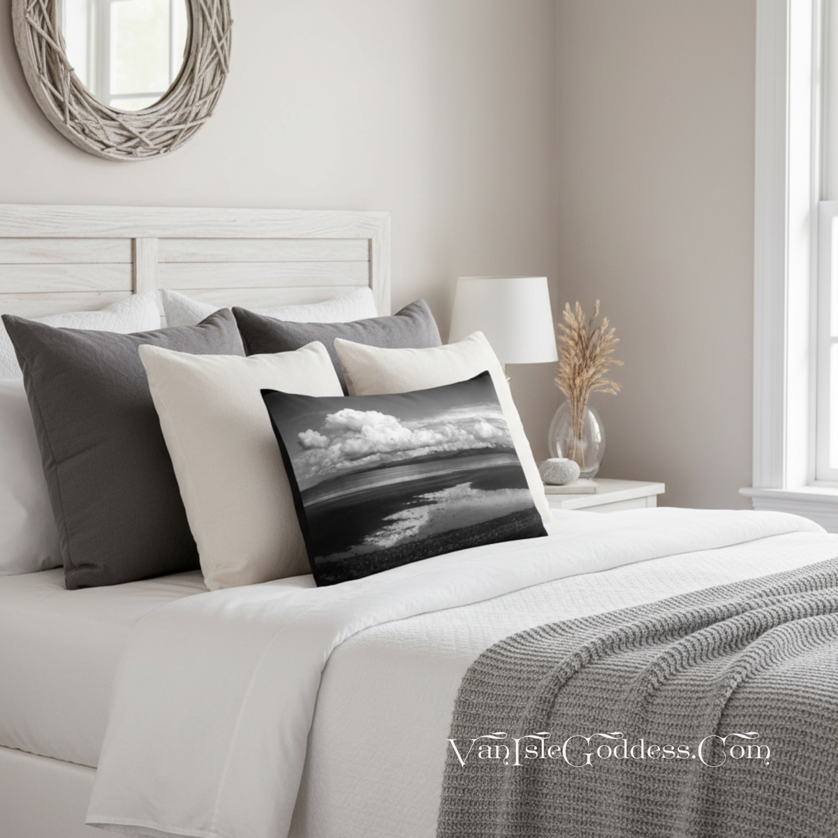 A styled bedroom scene with a bed dressed in white bedding and several pillows, including a square black-and-white landscape throw pillow. A gray knit throw blanket is draped across the foot of the bed, with a white nightstand, lamp, and window visible in the softly lit room.