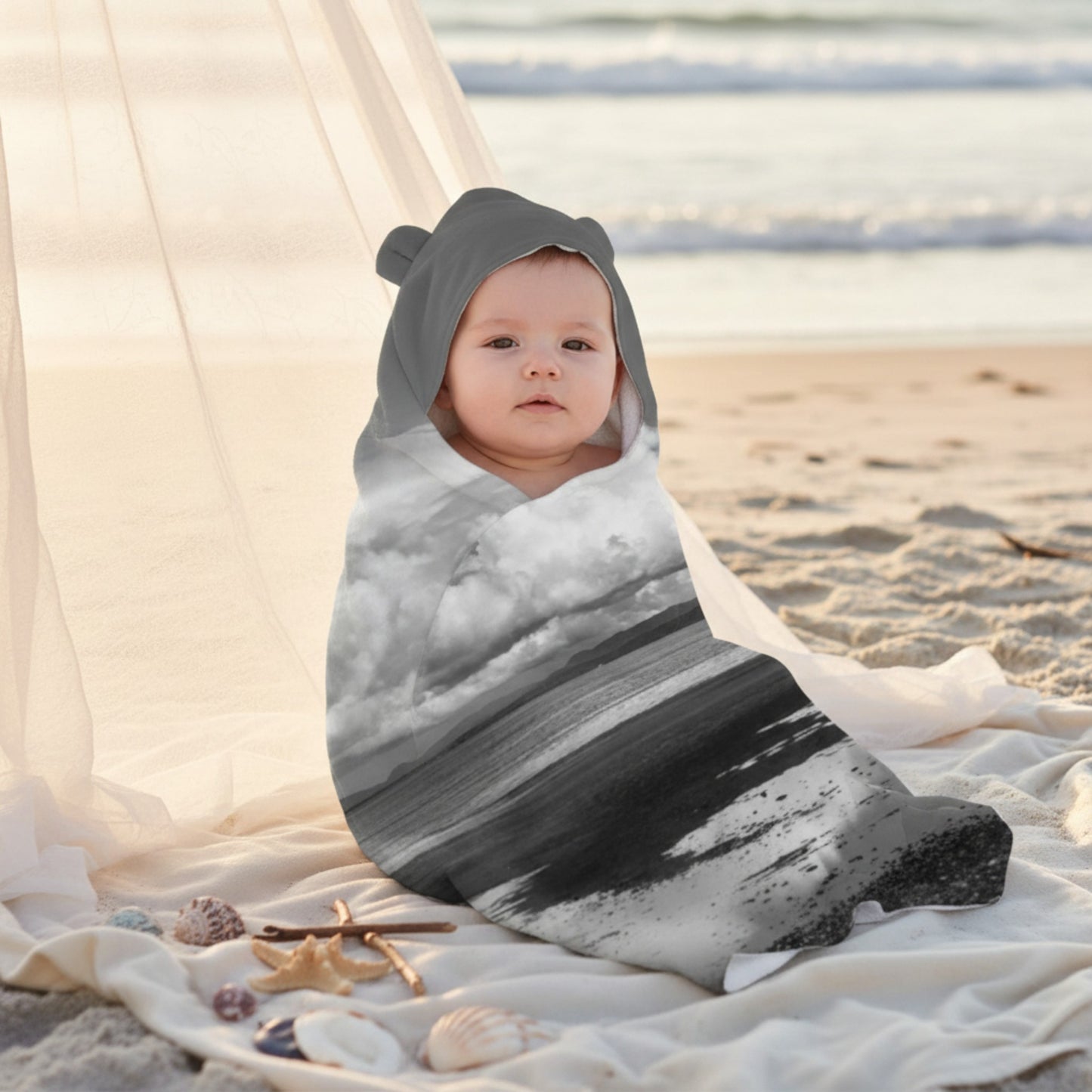 Baby wrapped in a gray and white striped swaddle on a sandy beach with water in the background