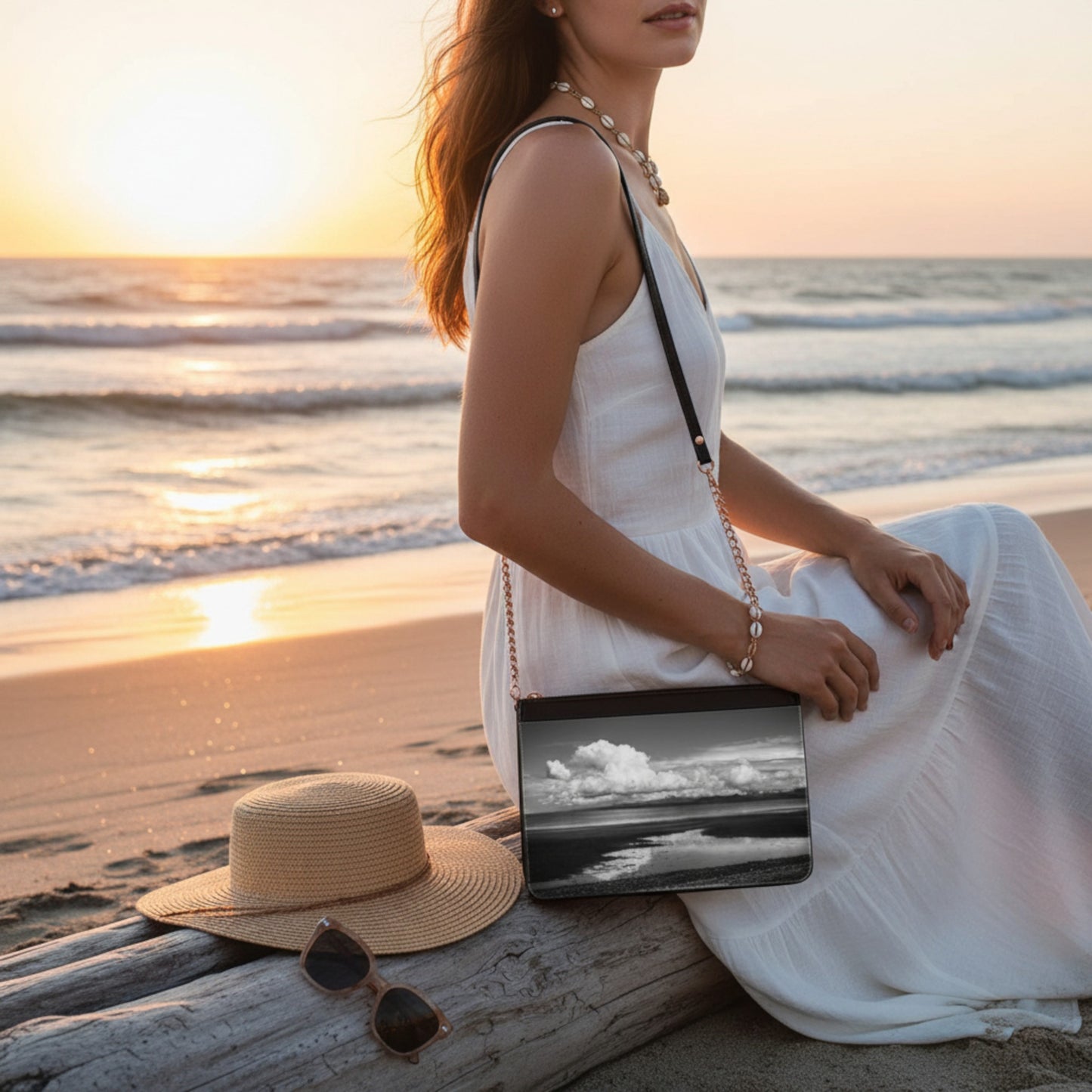 Woman in a white dress sitting on a beach at sunset with a hat and sunglasses beside her.