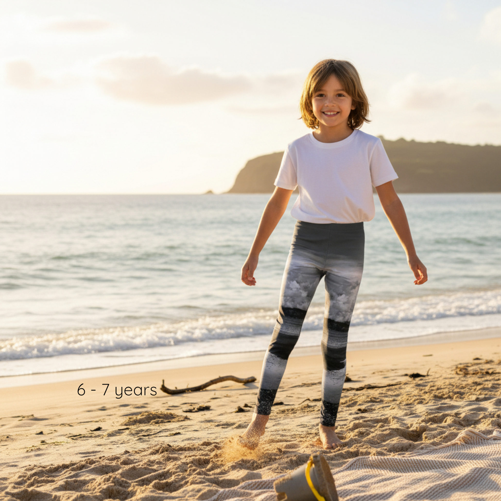 Child wearing a white t-shirt and patterned leggings standing on a beach with ocean and sunset in the background.