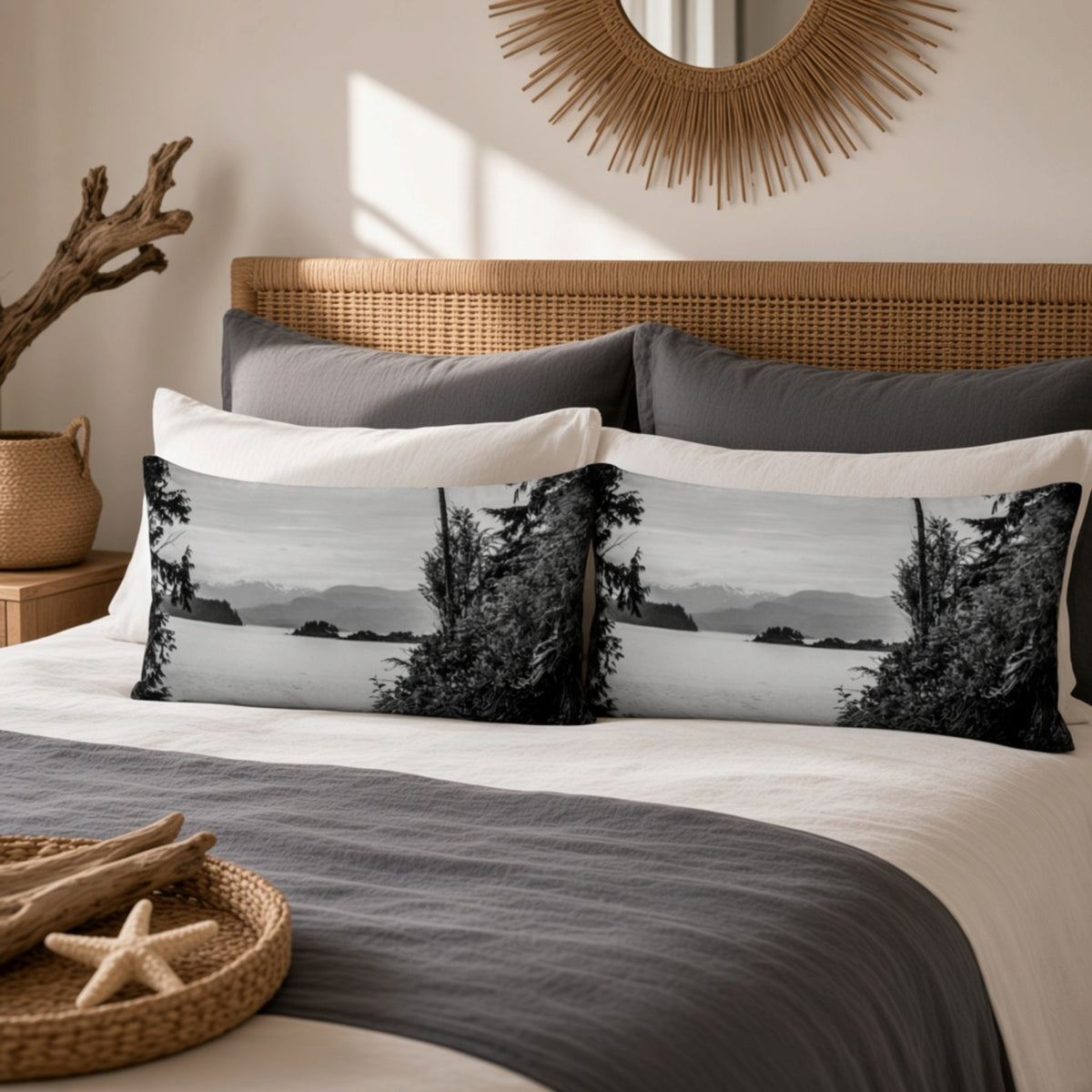 A styled bedroom scene with a bed made up with white and dark gray bedding and two long rectangular pillows featuring a black-and-white coastal landscape print with trees and distant hills. A wicker headboard, sunburst wall mirror, and decorative tray on the bed are visible.