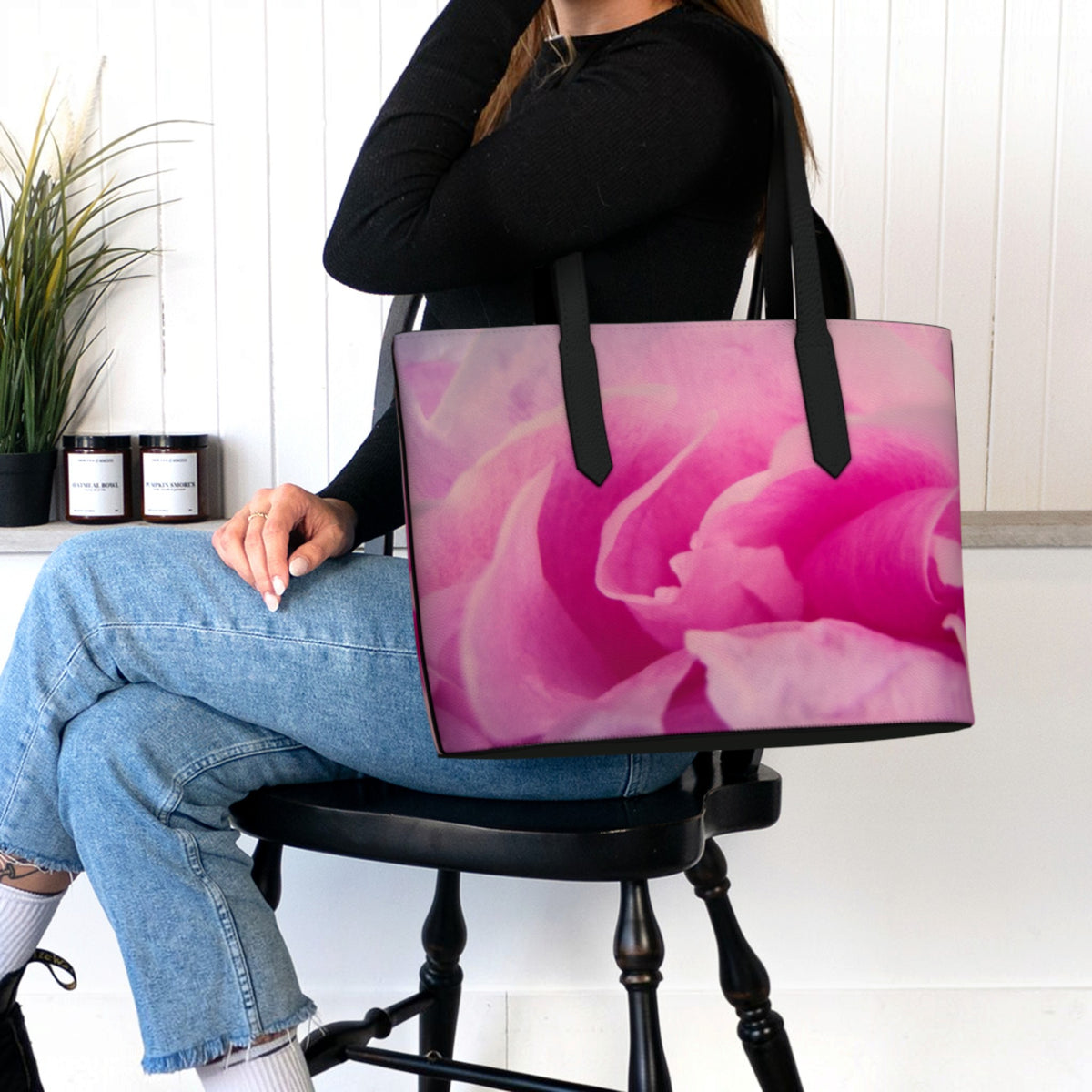 A person seated on a black stool holds a large tote bag featuring a pink rose print. The person is wearing a black long-sleeve top, light blue jeans, and white socks in a bright indoor setting with a white paneled wall, a plant, and small jars on a shelf.