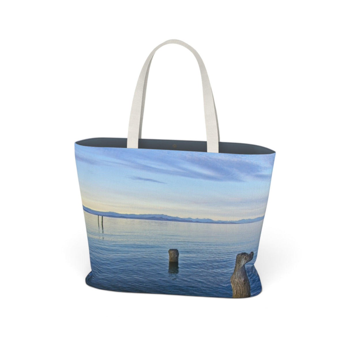 A tote bag featuring a scenic print of a calm lake with distant mountains under a blue sky, with white shoulder straps, shown on a plain white background.