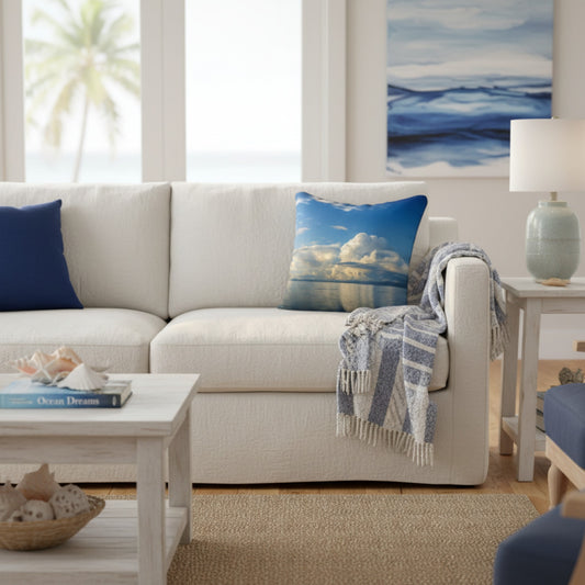 A styled living room scene featuring a white textured sofa with a blue and white cloud-patterned throw pillow, a navy blue accent pillow, and a blue-and-white fringed throw blanket. A coffee table, side table with a lamp, and a large blue ocean art print are visible in the background.