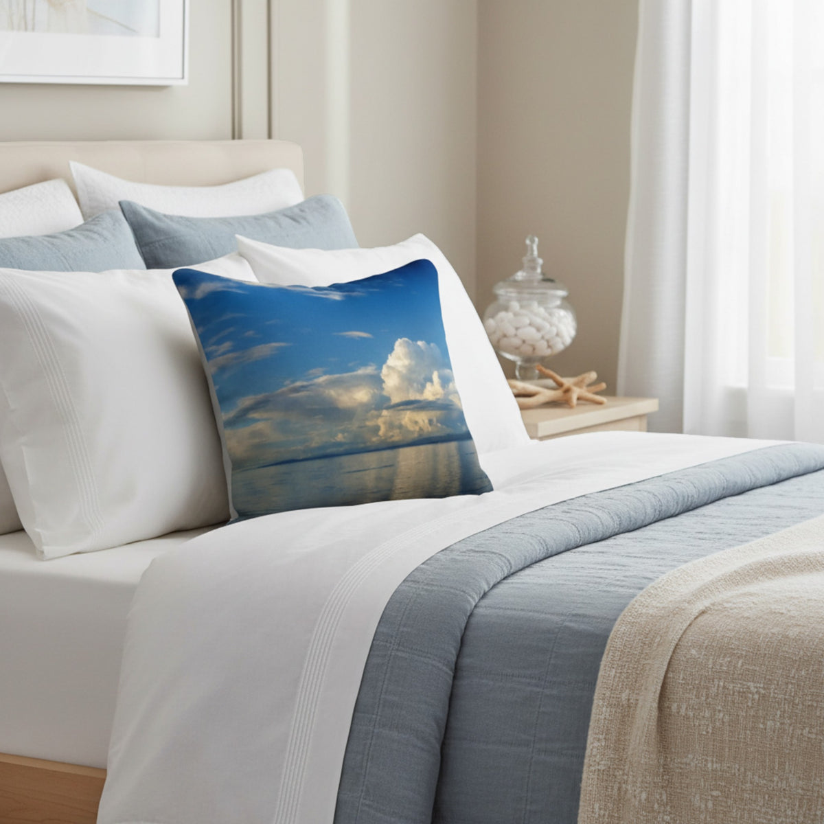 A square decorative throw pillow featuring a scenic blue sky and white clouds print, styled on a bed with white and blue bedding in a bright, modern bedroom.