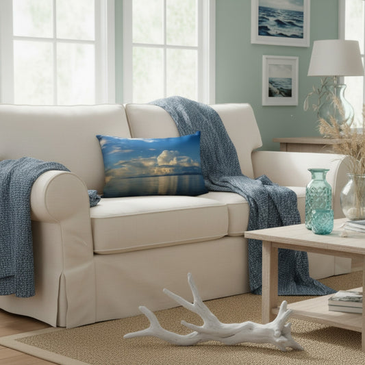 A cream slipcovered loveseat in a bright coastal-style living room, styled with a blue textured throw blanket and a square blue pillow featuring a sunset over water.