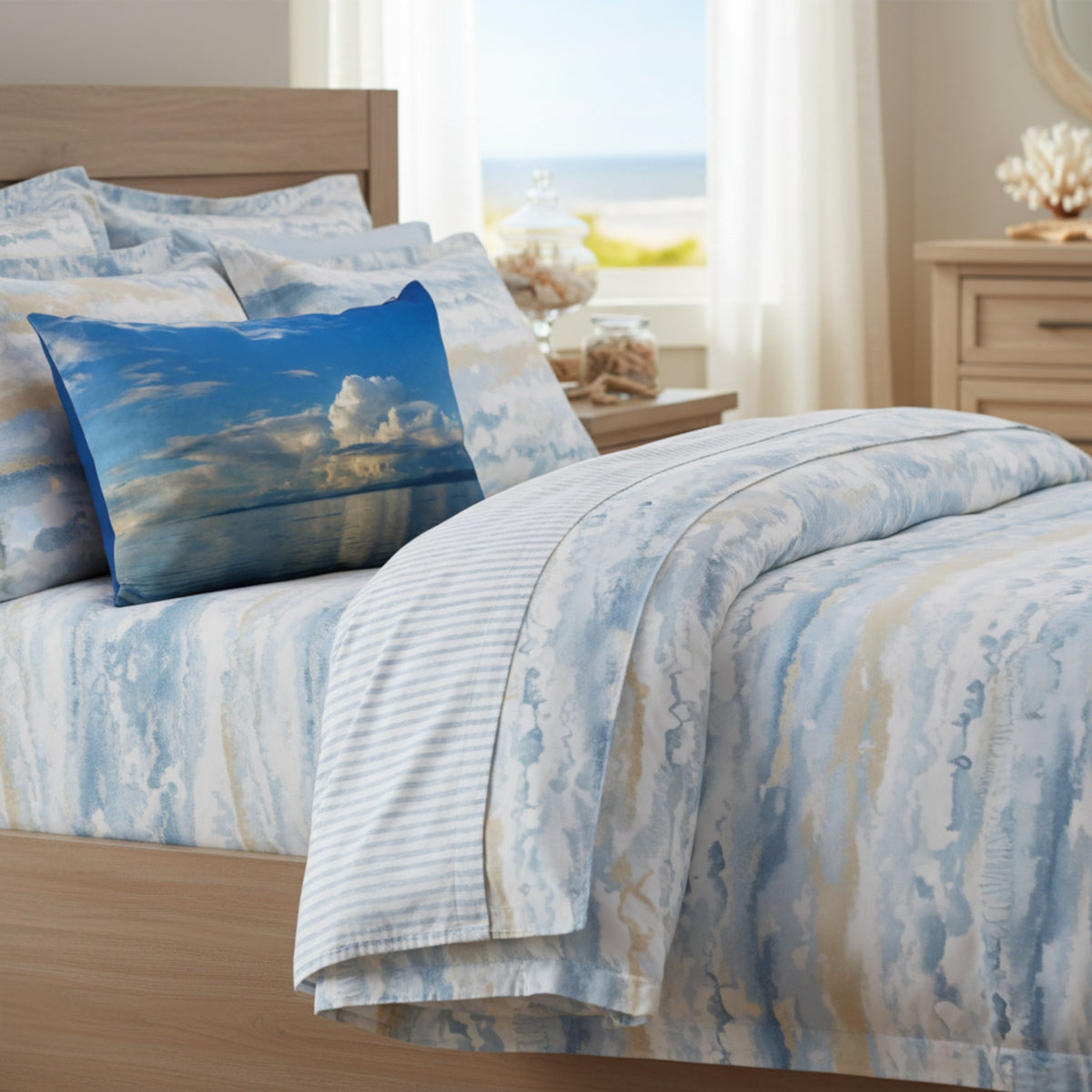 A bed dressed in a blue and white beach-inspired print bedding set, including a duvet cover, striped pillow shams, and a matching square throw pillow showing a sky and sea scene, styled in a bright bedroom.