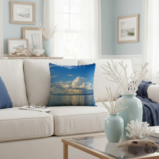 A square throw pillow featuring a scenic blue sky and white clouds over water, styled on a cream-colored sofa in a bright, coastal-themed living room.