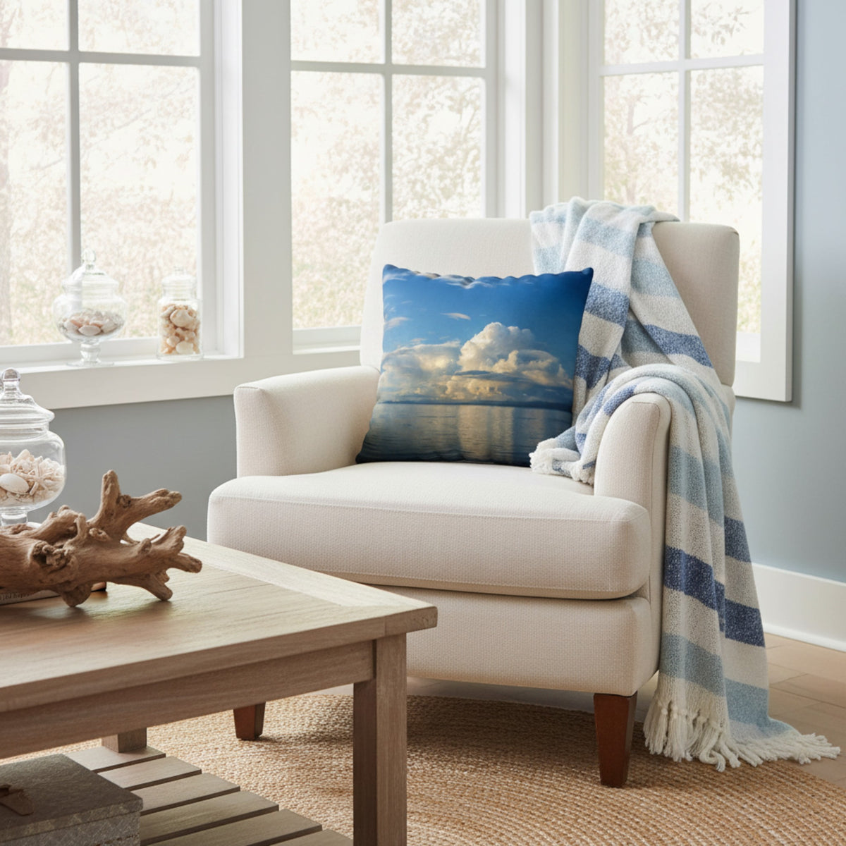 A square throw pillow featuring a scenic blue sky and white cloud reflection over water, styled on a cream armchair next to a blue and white striped throw blanket in a bright room.