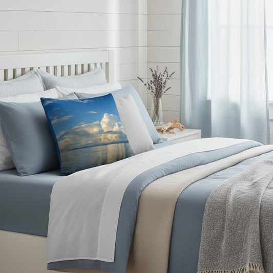 A styled bed in a bright bedroom setting with a blue and white bedding set, a beige throw blanket, and a decorative square pillow featuring a scenic sky and water print.