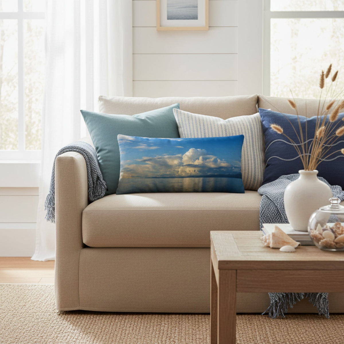 A beige sofa in a bright living room styled with several throw pillows, including a central rectangular pillow featuring a blue sky and white cloud landscape print.