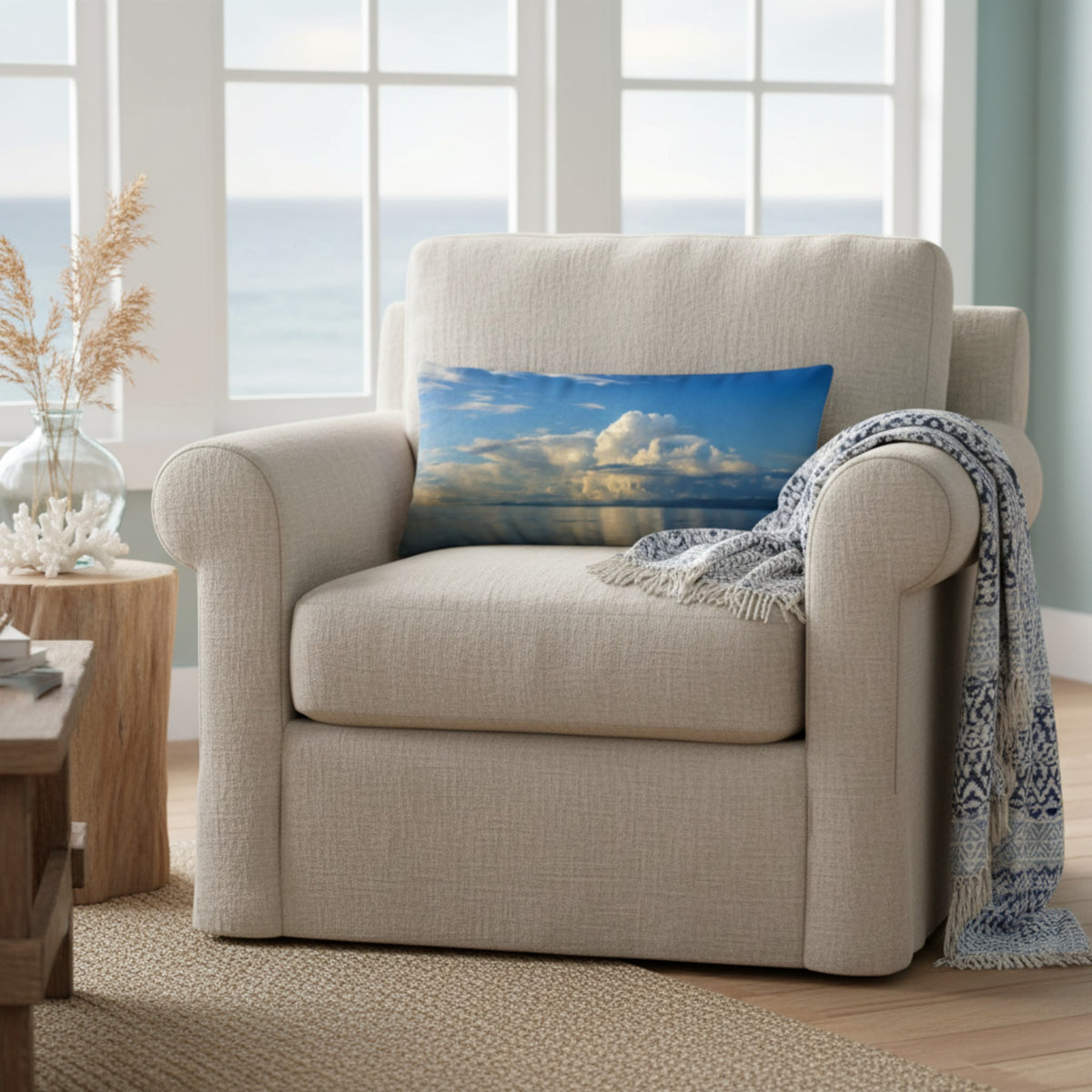 A beige upholstered armchair in a bright living room, styled with a square throw pillow showing a blue sky and white clouds, and a blue-and-white fringed throw blanket draped over the arm.