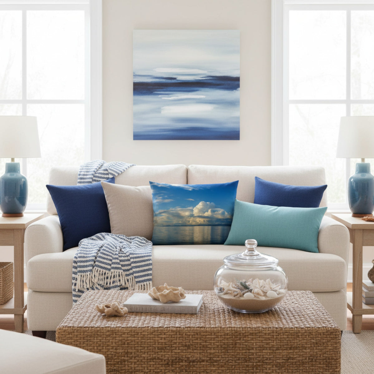 A coastal-style abstract canvas print in shades of blue and white hangs on a beige wall above a cream-colored sofa styled with blue and teal throw pillows in a bright, modern living room.