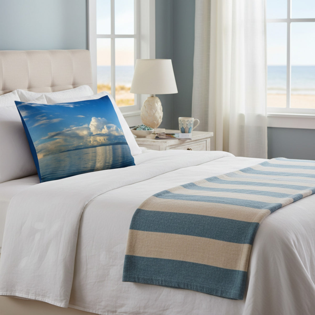 A beach-themed pillow sham with a blue sky and ocean print styled on a bed with white linens and a blue and beige striped throw blanket in a bright, modern bedroom.