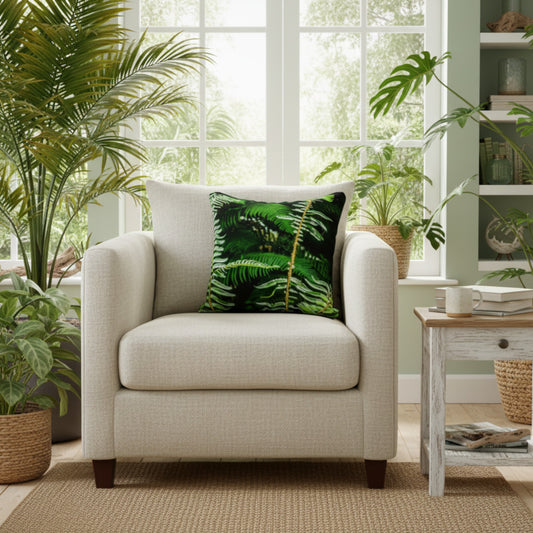 A light beige upholstered armchair in a bright living room setting, styled with a dark green palm leaf patterned throw pillow. The chair is positioned on a woven jute rug next to a distressed white side table with a mug, with potted plants and a window in the background.