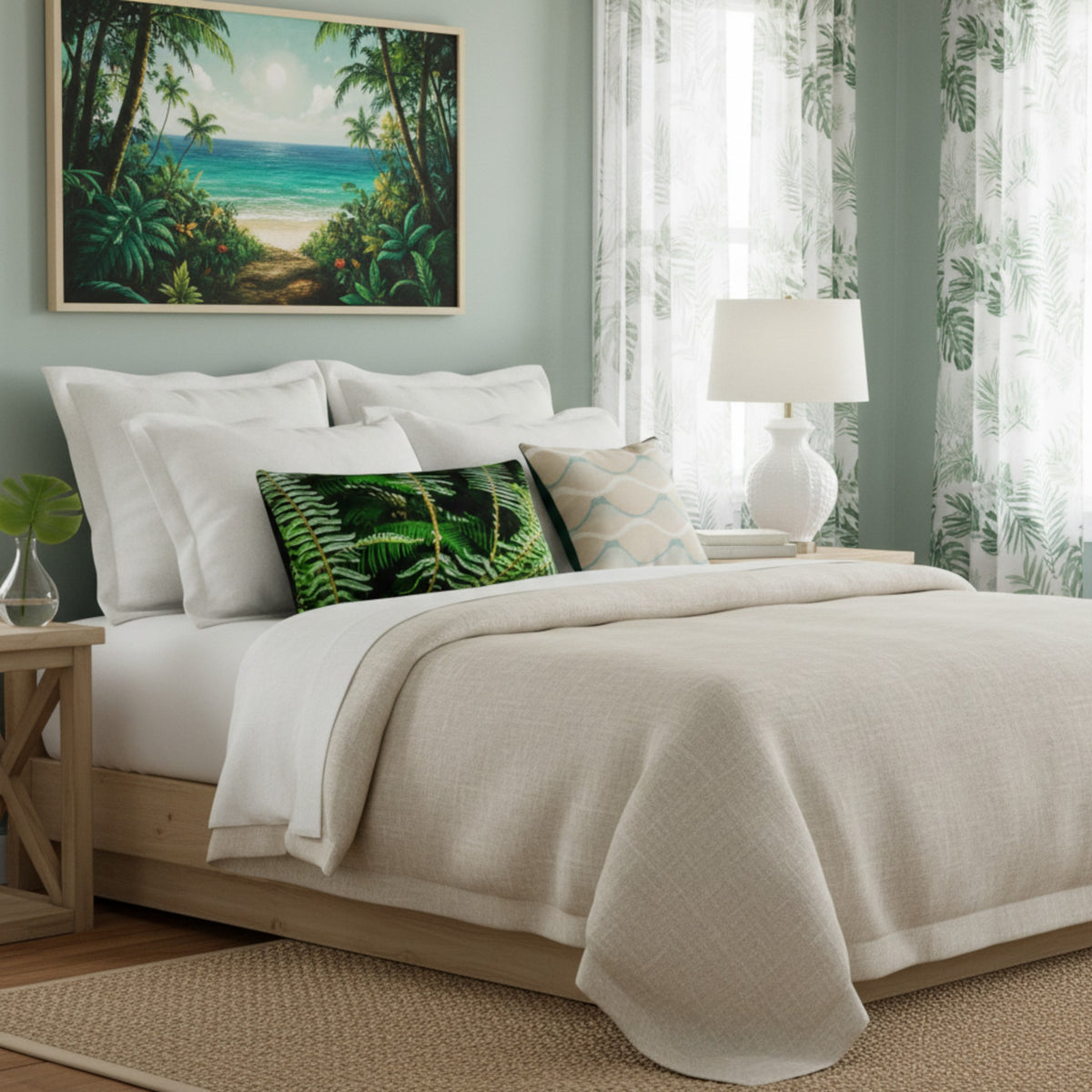 A styled bedroom scene with a bed dressed in white bedding and a textured cream throw blanket, accented with green and beige decorative pillows. A tropical beach painting hangs on the wall above, with sheer green leaf-print curtains and a white table lamp on a nightstand.