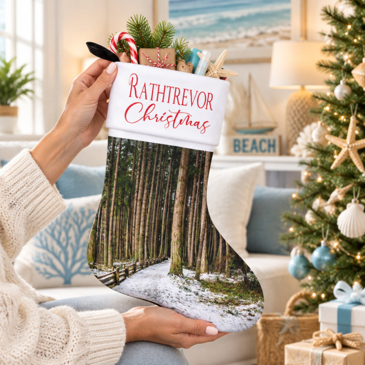 A person holds a personalized Christmas stocking in a cozy living room setting. The stocking features a white top with red text reading 'RATHTREVOR Christmas' and a forest scene printed below.