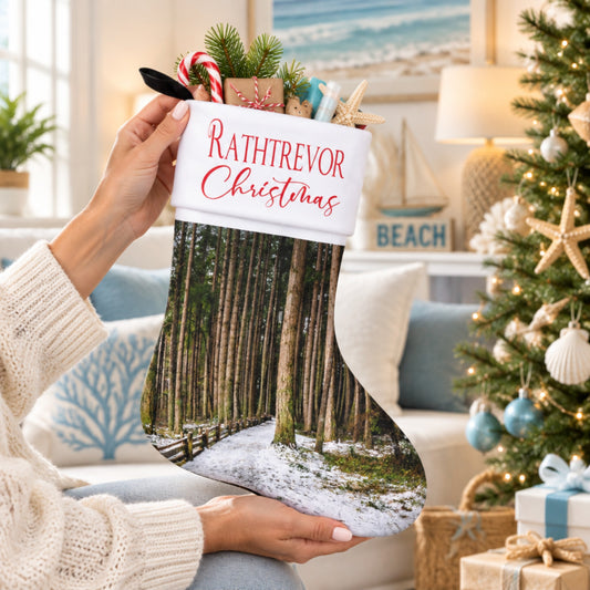 A person holds a personalized Christmas stocking in a cozy living room setting. The stocking features a white top with red text reading 'RATHTREVOR Christmas' and a forest scene printed below.