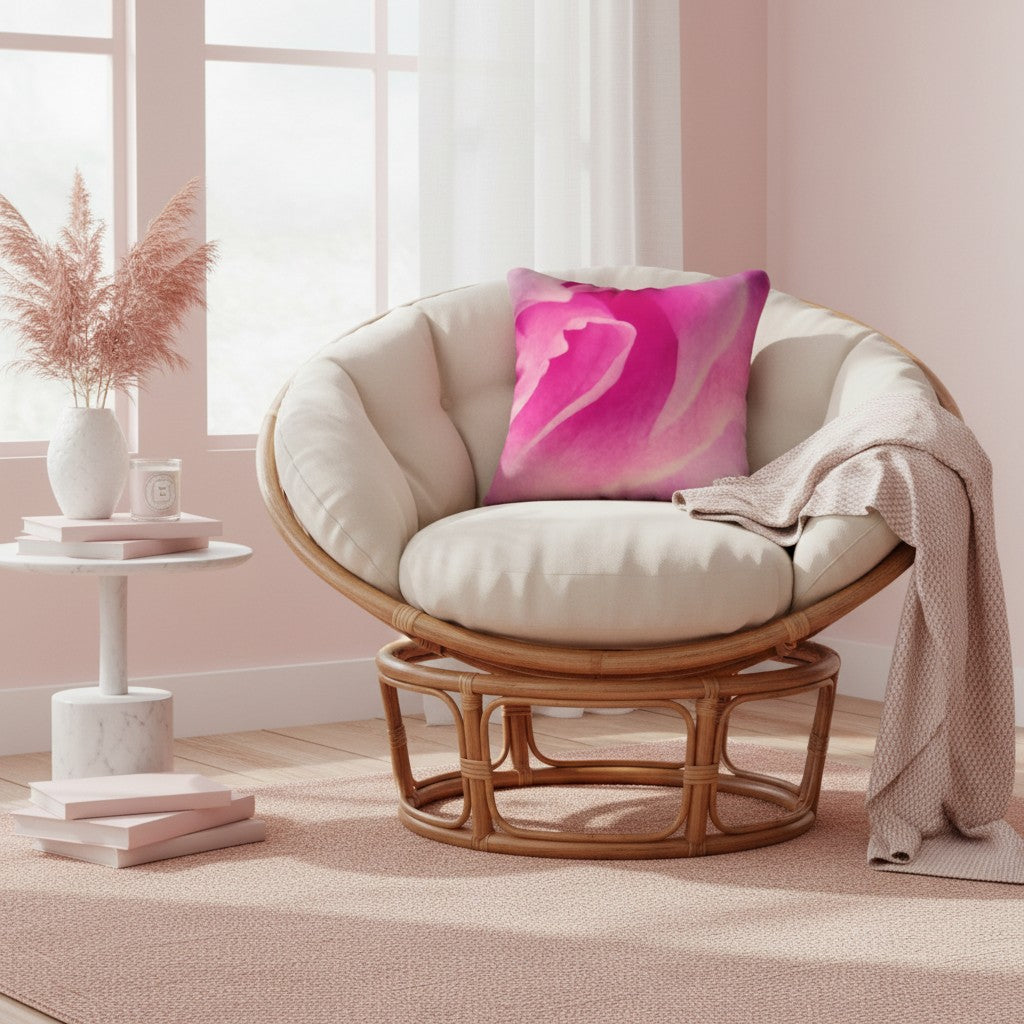 A round rattan swivel chair with cream cushions and a bright pink abstract throw pillow, styled in a soft pink room with a draped blanket, a small side table, and a window.