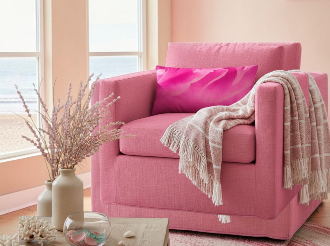 A pink upholstered armchair in a bright room by large windows, styled with a bright pink pillow, a beige fringed throw blanket, and a vase of dried stems on a table in the foreground.