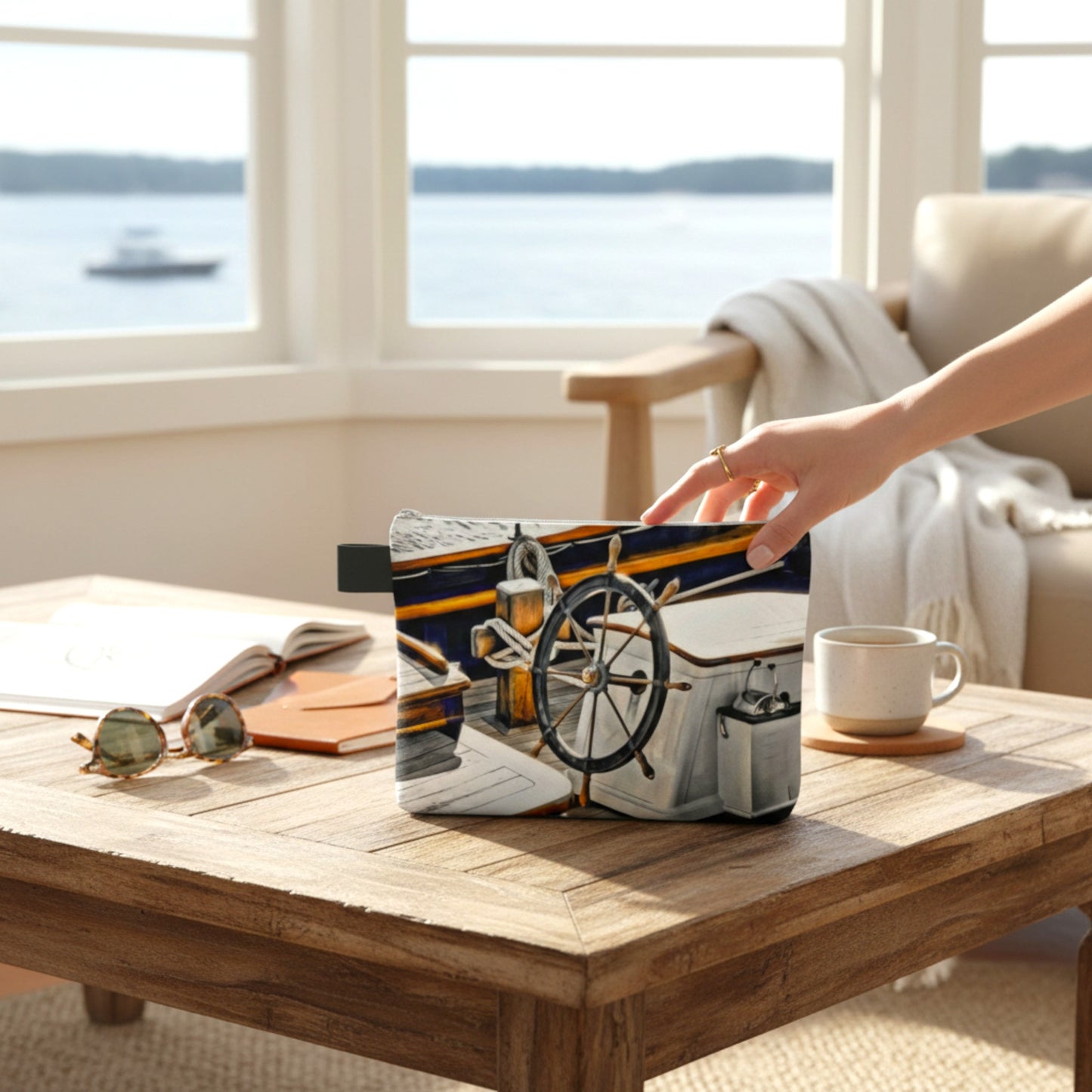 Hand holding a tote bag with a ship design on a wooden table by a window with a view of water.